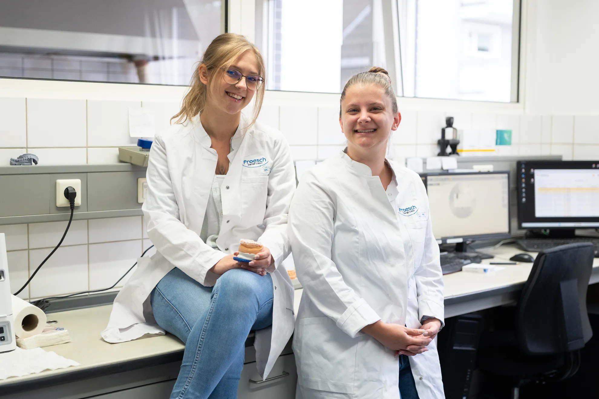 Zwei Frauen in weißen Laborkitteln stehen in einem Labor. Eine sitzt auf einem Tisch und hält ein Zahnmodell, während die andere steht.