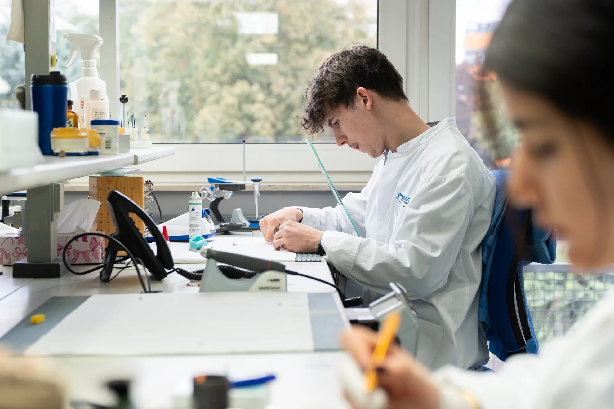 Ein Mann arbeitet konzentriert am Tisch, umgeben von verschiedenen Geräten und Flaschen; im Vordergrund ist eine weitere Person unscharf